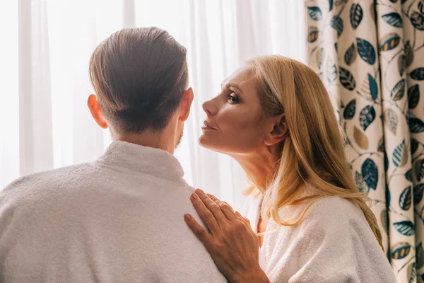 Vista de perfil de mujer madura capaz de besar a su marido con albornoz en la habitación de hotel - foto de stock
