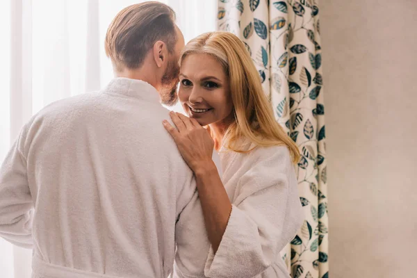 Feliz pareja madura en albornoces abrazos y la mujer sonriendo a la cámara en la habitación del hotel - foto de stock