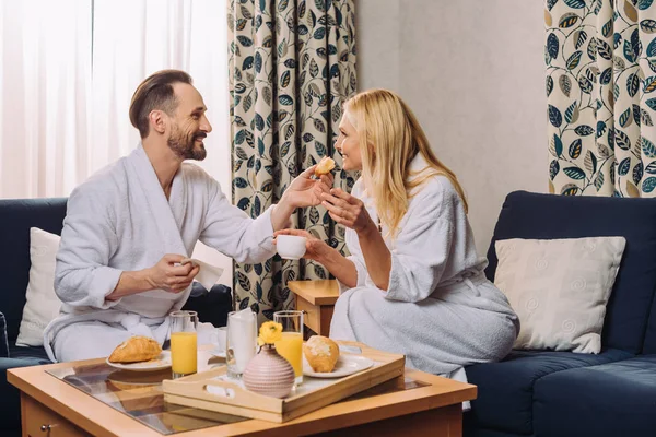 Feliz hombre maduro en albornoz alimentar a su esposa con pastelería durante el desayuno en la habitación de hotel - foto de stock