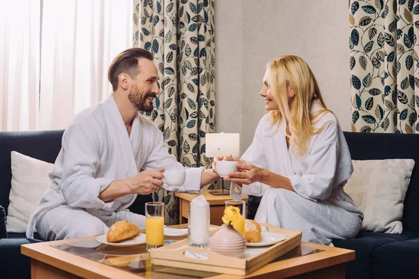 Happy middle aged couple in bathrobes drinking coffee during breakfast in hotel room — Stock Photo
