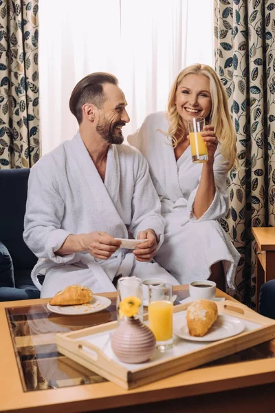 Feliz pareja de mediana edad en albornoces desayunando juntos en la habitación del hotel - foto de stock