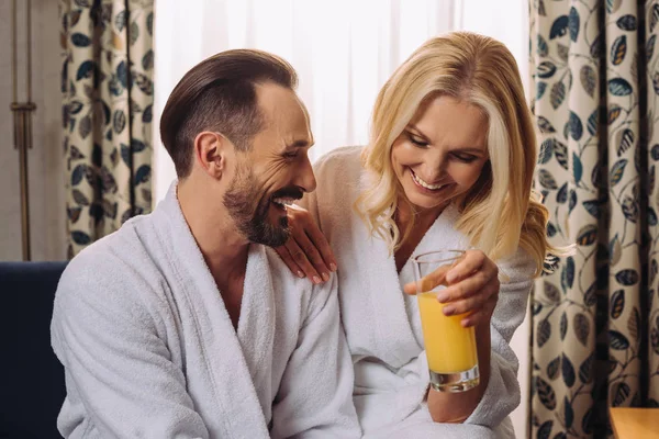 Feliz pareja de mediana edad en albornoces beber jugo durante el desayuno en la habitación del hotel - foto de stock