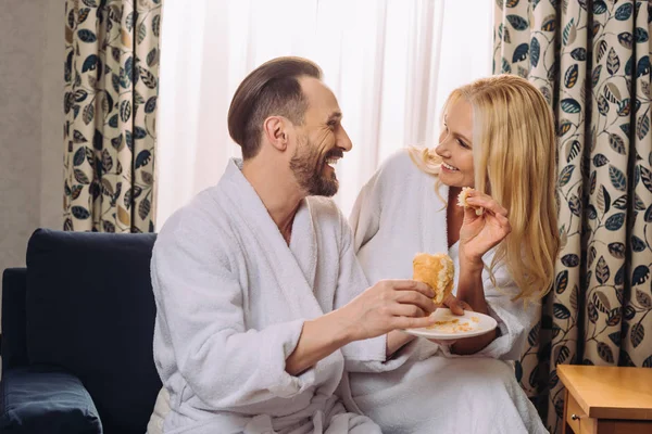 Счастливая взрослая пара в халатах, вместе завтракающая выпечкой в номере отеля — стоковое фото