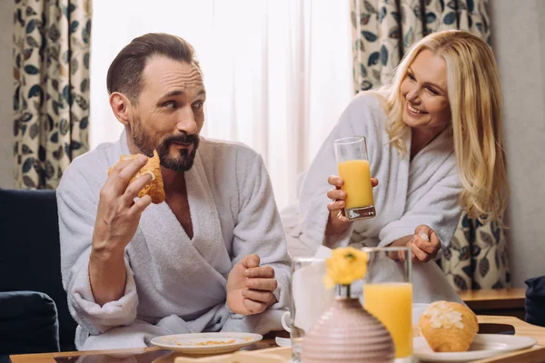 Sourire couple d'âge moyen boire du jus et manger de la pâtisserie pendant le petit déjeuner dans la chambre d'hôtel — Photo de stock