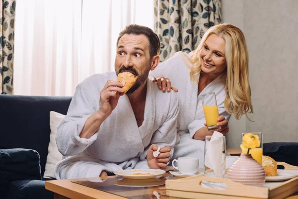 Feliz pareja adulta beber jugo y comer pastelería durante el desayuno en la habitación de hotel - foto de stock