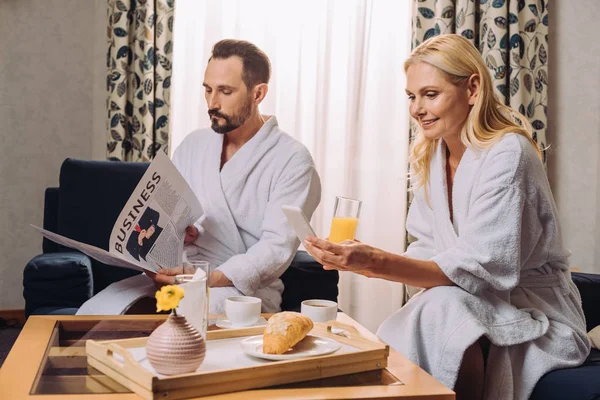 Couple mature souriant en peignoirs tenant journal et smartphone tout en prenant le petit déjeuner dans la chambre d'hôtel — Photo de stock