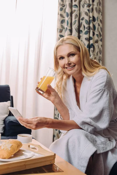 Mujer madura en albornoz sonriendo a la cámara mientras bebe jugo y usando el teléfono inteligente en la habitación del hotel - foto de stock