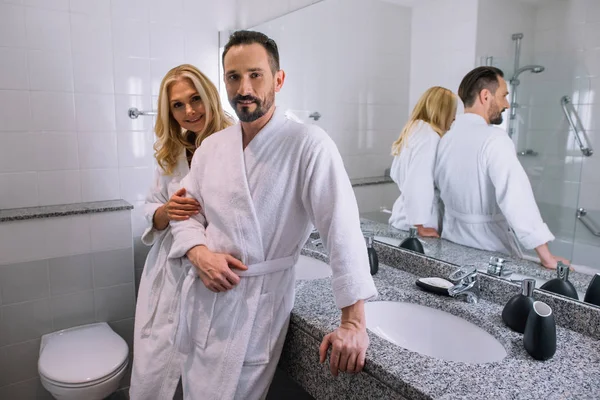 Щаслива зріла пара в халатах посміхається на камеру у ванній кімнаті готелю — стокове фото