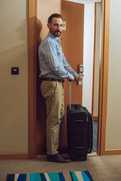 Bärtiger Mann mit Koffer blickt in Kamera beim Betreten des Hotelzimmers — Stockfoto