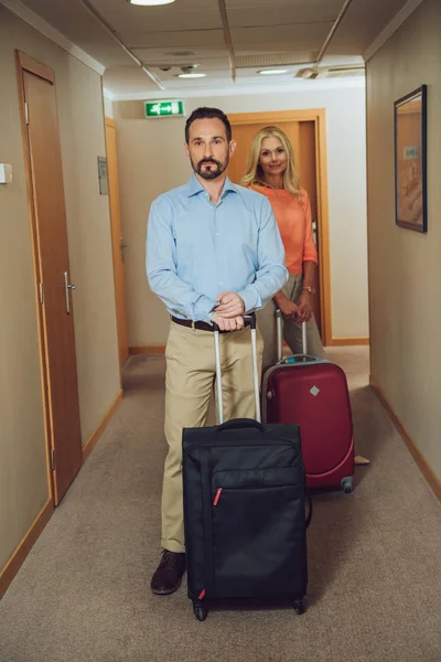 Mature couple with suitcases looking at camera while standing in hotel corridor — Stock Photo