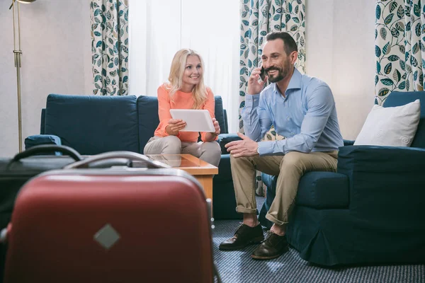 Pareja madura sonriente usando teléfono inteligente y tableta digital mientras se sienta con maletas en la habitación del hotel - foto de stock