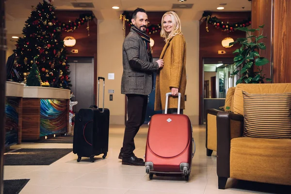 Middle aged couple standing with suitcases and smiling at camera in hotel hallway at christmas — Stock Photo