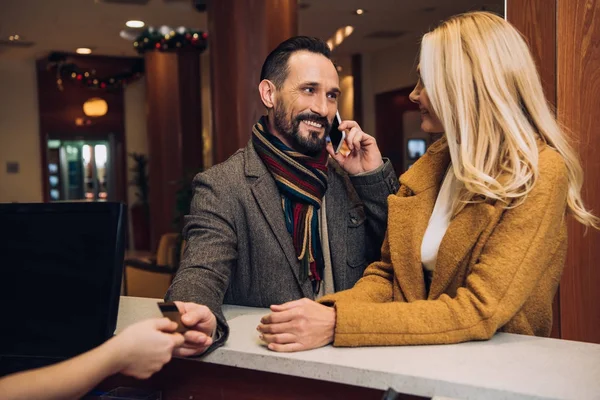 Bela mulher madura olhando para o marido falando no smartphone e dando cartão de crédito para recepcionista no hotel — Fotografia de Stock