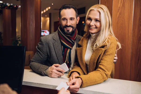 Sorrindo casal maduro segurando smartphone e cartão na recepção no hotel — Fotografia de Stock