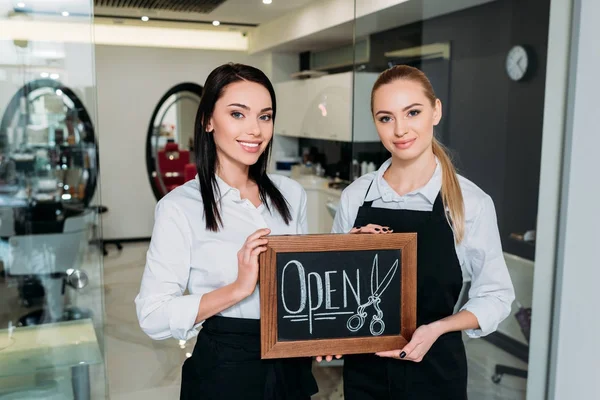 Beautiful hairdressers standing with signboard open — Stock Photo