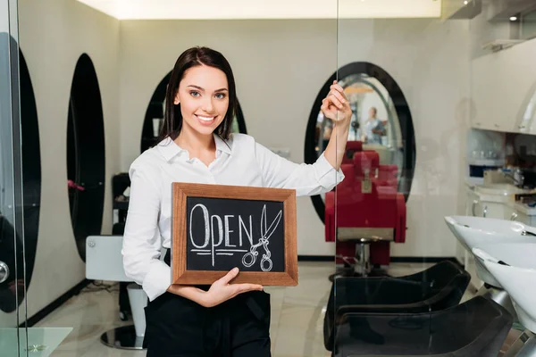 Sonriente dueño de peluquería de pie con el cartel abierto y apoyado en la puerta de cristal - foto de stock