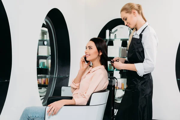 Mulher falando por smartphone enquanto cabeleireiro corte de cabelo — Fotografia de Stock