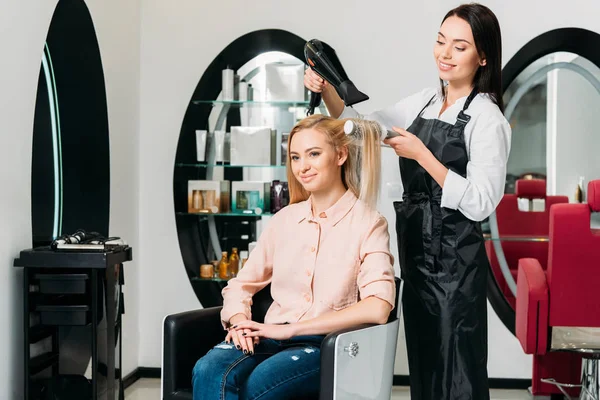 Peluquería sonriente secar el pelo del cliente en el salón - foto de stock