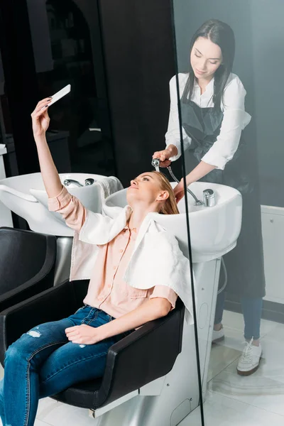 Beautiful woman taking selfie while hairdresser washing her hair at salon — Stock Photo