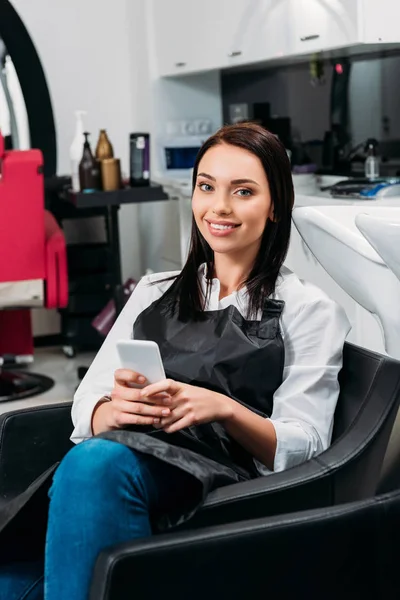 Friseur sitzt bei der Arbeit im Stuhl und hält Smartphone in der Hand — Stockfoto