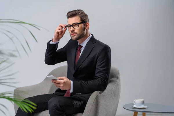 Businessman sitting in armchair with smartphone and looking away — Stock Photo