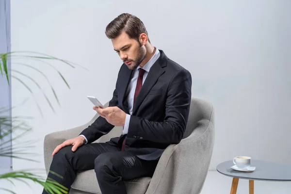 Hombre de negocios sentado en sillón y mirando el teléfono inteligente - foto de stock