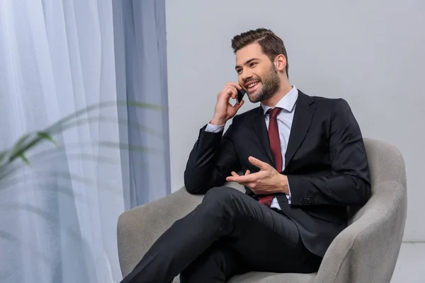 Smiling businessman sitting in armchair and talking by smartphone — Stock Photo