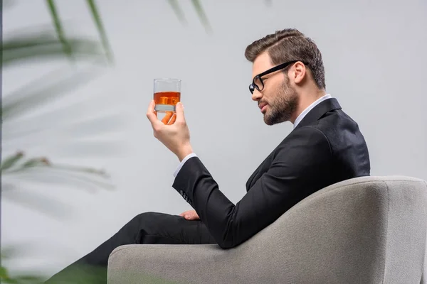 Hombre de negocios sentado en sillón y mirando un vaso de whisky - foto de stock
