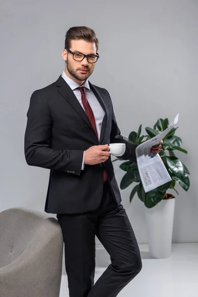 Handsome businessman holding cup of coffee and newspaper and looking at camera — Stock Photo