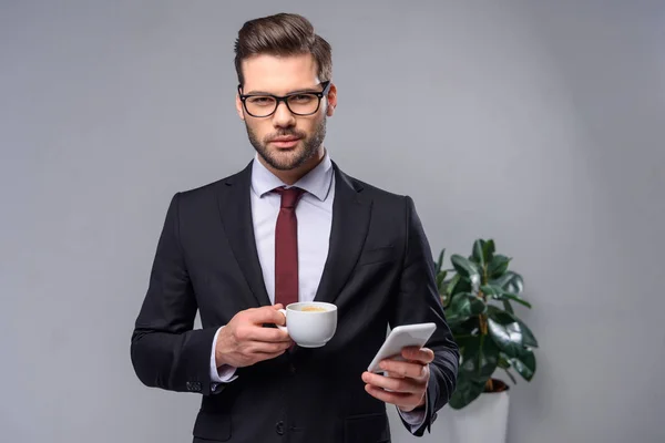 Homme d'affaires sérieux tenant smartphone et tasse de café et regardant la caméra — Photo de stock