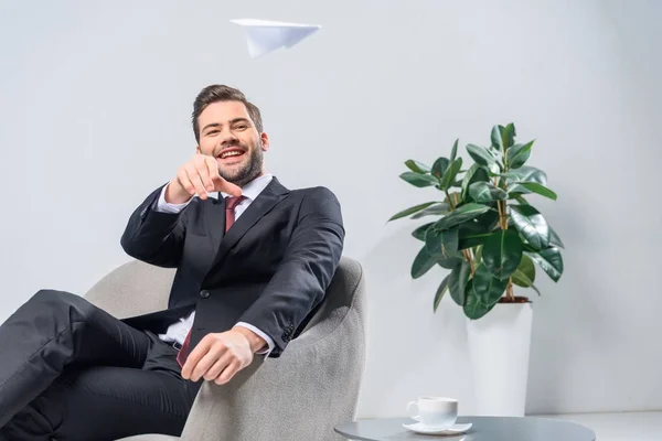 Sonriente hombre de negocios sentado en sillón y arrojando avión de papel en la oficina - foto de stock