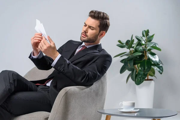 Hombre de negocios serio haciendo avión de papel mientras está sentado en el sillón - foto de stock