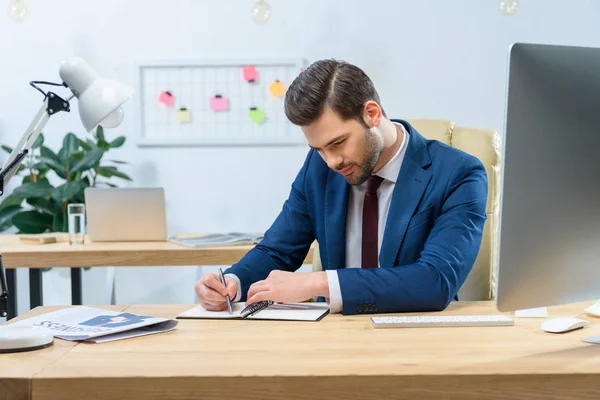 Gutaussehender Geschäftsmann schreibt am Tisch etwas ins Notizbuch — Stockfoto