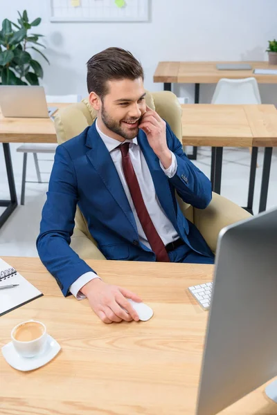 Sonriente hombre de negocios hablando por teléfono inteligente y mirando a la computadora - foto de stock