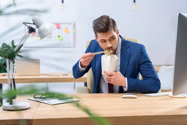 Empresário comer macarrão e olhando para o computador — Fotografia de Stock