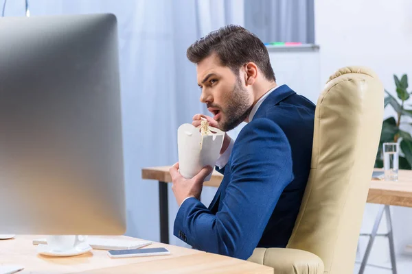 Hungry businessman eating noodles and looking at computer — Stock Photo