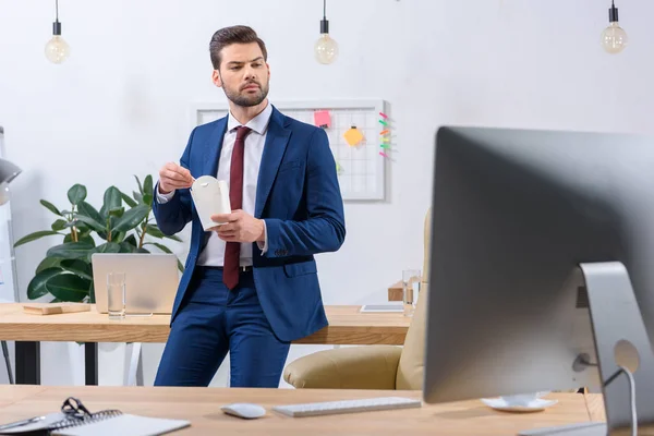 Seriöser Geschäftsmann hält Papierschachtel mit Nudeln in der Hand und schaut auf den Computer — Stockfoto