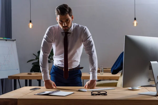 Hombre de negocios serio de pie y apoyado en la mesa de trabajo - foto de stock