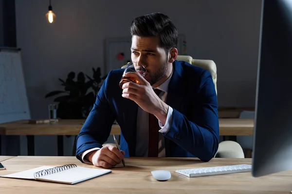 Businessman drinking whiskey in dark office and looking away — Stock Photo