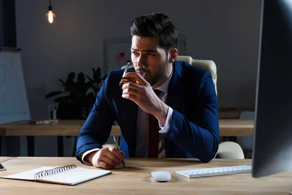 Hombre de negocios bebiendo whisky en la oficina oscura y mirando hacia otro lado - foto de stock