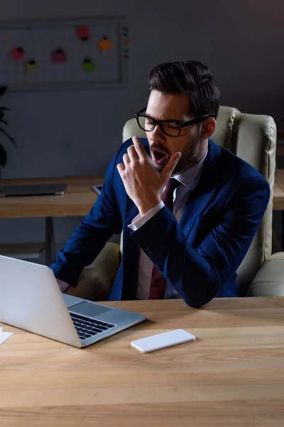 Бизнесмен работает за ноутбуком в офисе и зевает — стоковое фото