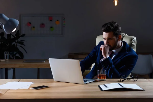 Задумчивый бизнесмен сидит в темном офисе и смотрит на ноутбук — стоковое фото