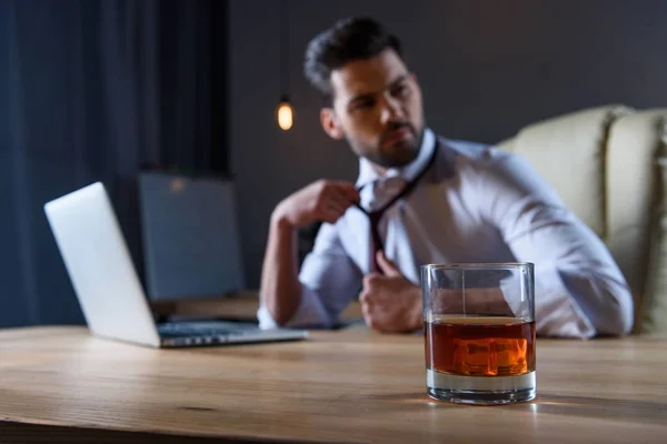 Empresário cansado soltar gravata e sentado à mesa com um copo de uísque em primeiro plano — Fotografia de Stock