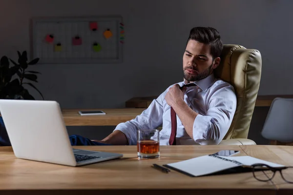 Tired businessman loosen tie and sitting at table with glass of whiskey — Stock Photo