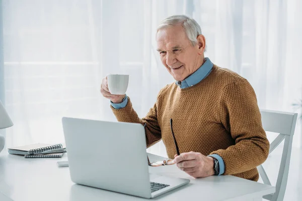 Hombre seguro de edad que trabaja en el ordenador portátil y la celebración de taza de café - foto de stock