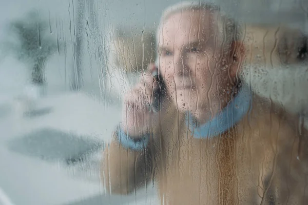 Detrás de la vista de cristal lluvioso de un hombre mayor perturbado haciendo una llamada telefónica - foto de stock