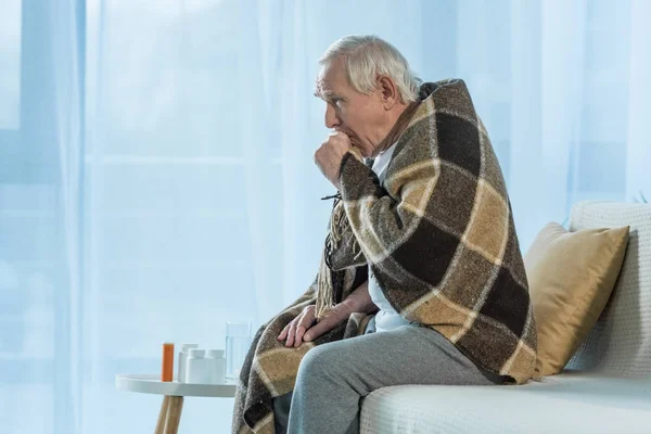 Senior chilled man covered in plaid coughs while sitting sofa in room with medications on table — Stock Photo