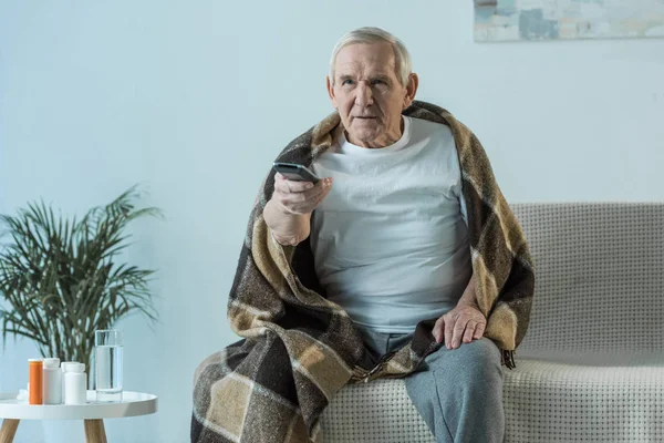 Enfermo hombre mayor cubierto de cuadros mira la televisión en la habitación con medicamentos en la mesa - foto de stock