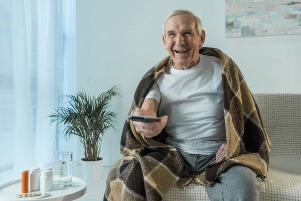 Homme âgé couvert de plaid regarde la télévision pendant la maladie dans la chambre avec des médicaments sur la table — Photo de stock