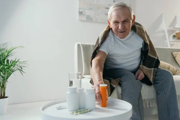 Senior sick man covered in plaid takes medications from table — Stock Photo